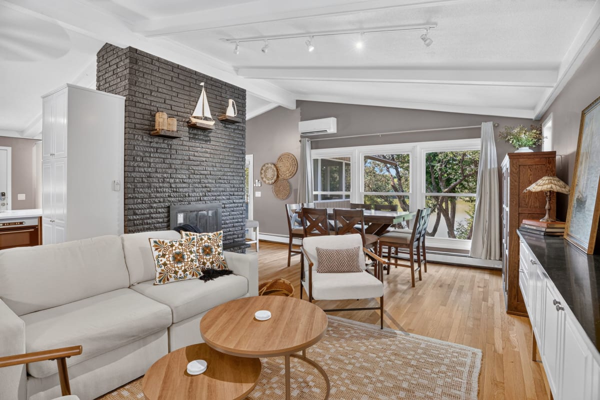 Unwind in your open-concept living and dining space, where a striking brick fireplace (decorative only) anchors the room and large windows frame lush greenery and a view of the lake just outside.