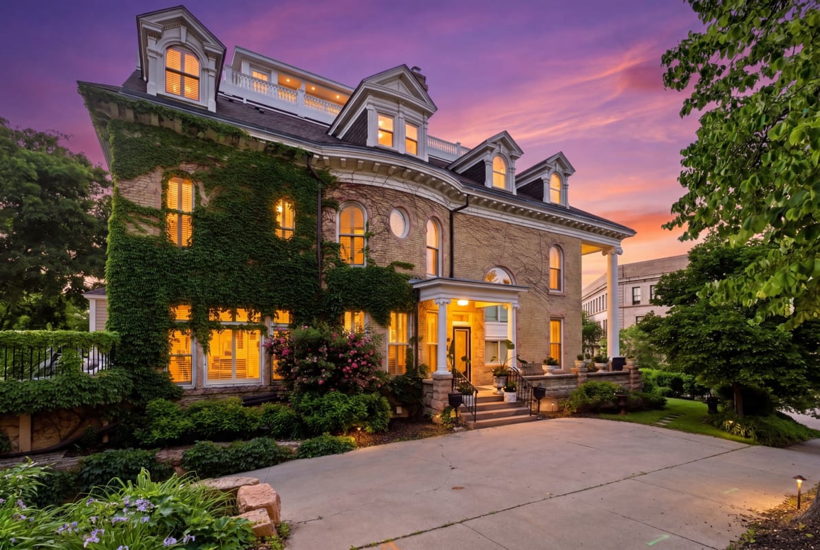 Ivy-draped Victorian elegance welcomes arrivals beneath a stunning purple-pink sunset. The historic brick facade glows with warm interior lighting, framed by mature gardens and a welcoming entrance.