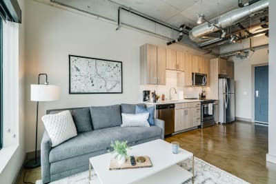 A modern apartment with a living area and kitchen, showcasing a gray sofa, floor lamp, and stainless steel appliances.