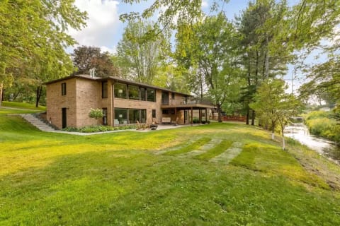 A two-story brick house surrounded by trees and a well-kept lawn, near a stream.
