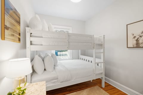 A bright bedroom with a white bunk bed and warm lighting, featuring decorative pillows and a friendly animal print on the wall.