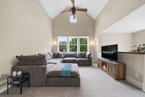 A modern living room with a large gray sectional sofa, a flat-screen TV, and tall windows letting in sunlight.