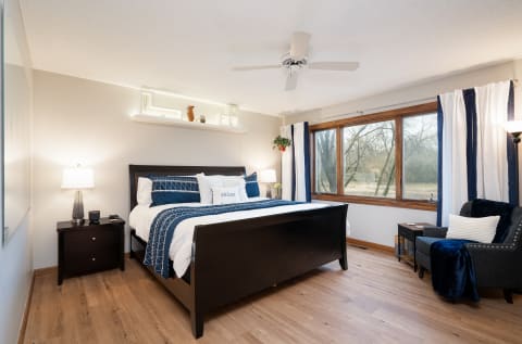 A well-decorated bedroom with a dark wood bed, pillows, a nightstand with lamp, and a large window overlooking trees.