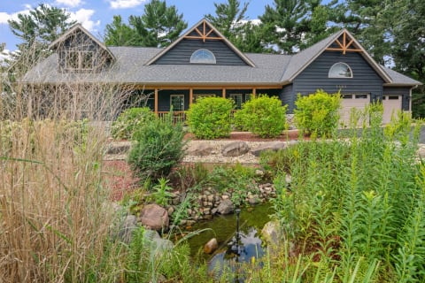 A modern dark-colored house with a porch, surrounded by gardens and a small pond.