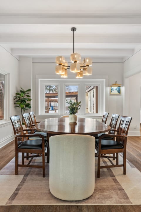 A modern dining room featuring a round wooden table, elegant chairs, and a striking chandelier, with large windows and a potted plant.