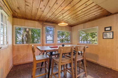 Cozy cabin dining room with wooden table and chairs, looking out onto greenery and water.