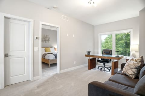 Bright living area with a sofa and desk bathed in natural light, leading to a bedroom.