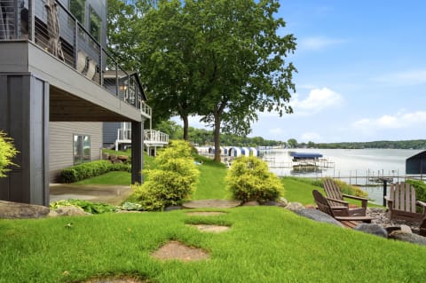 Lakeside property with lush grass, patio chairs, and boat docks under a bright blue sky.