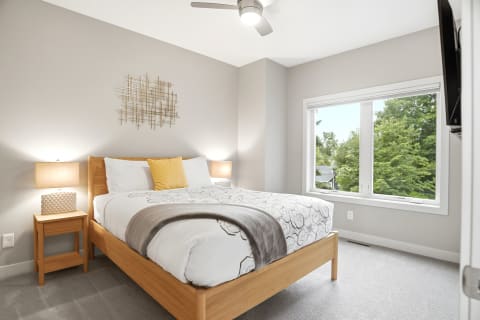 A modern bedroom with a queen bed, textured lamp, and large windows showcasing trees outside.
