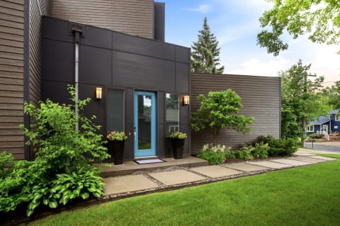 A contemporary home entrance with a turquoise door and dark paneling, surrounded by lush greenery.