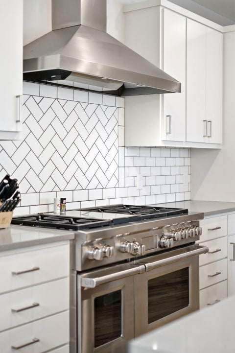 Modern kitchen with stainless steel gas range, herringbone tile backsplash, and white cabinetry.
