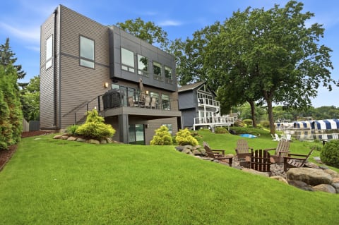 Modern lakeside home with a spacious deck, manicured lawn, and fire pit seating surrounded by lush greenery.