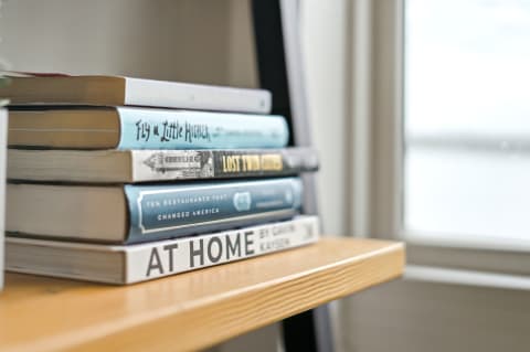 A stack of books in a cozy, light-filled setting on a natural wood shelf.
