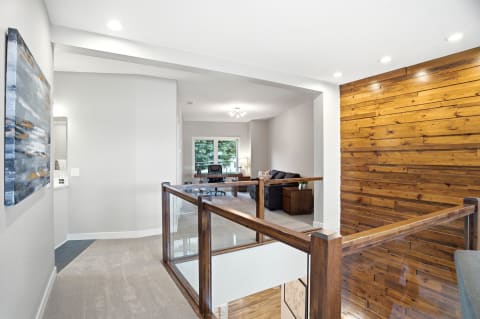 Open hallway with wood accent wall and glass railing, leading to a well-lit living area with modern furniture.