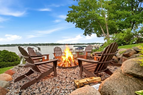 A lakeside fire pit surrounded by Adirondack chairs on pebbled ground, with trees and water in the background.