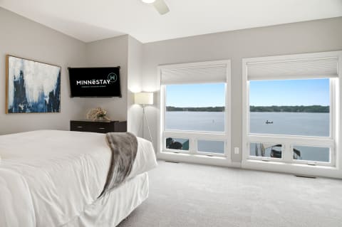 A serene bedroom with large windows showing a lake view, modern furnishings, and a cozy bed.