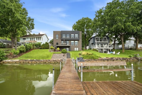 Modern lakeside home with spacious dock, featuring manicured lawns and multiple residences in a tranquil setting.