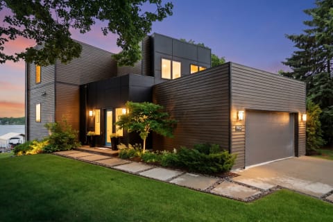 A contemporary lakeside home with black and wood panels, illuminated entrance, and landscaped path, under a twilight sky.