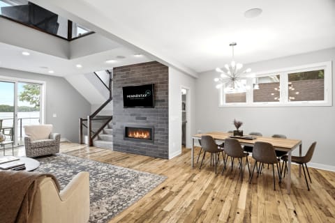 A spacious living room and dining area with a fireplace, wooden floors, and large windows overlooking outdoor scenery.