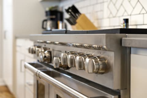 A professional-grade stainless steel oven range with polished knobs and a geometric tiled backsplash in a modern kitchen setting.
