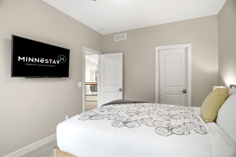 A tranquil bedroom with a plush bed, mounted TV displaying MINNESTAY, and neutral-colored walls.