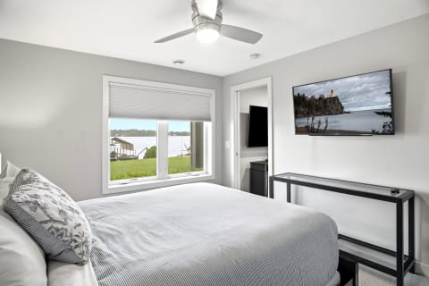A cozy bedroom with a queen bed, flat-screen TV, and a large window overlooking a scenic lake.