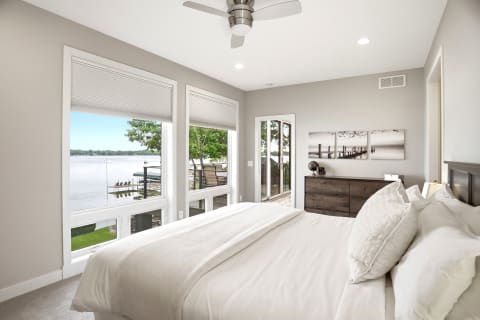 A bright bedroom with large windows overlooking a peaceful lake, featuring a cozy bed and modern decor.