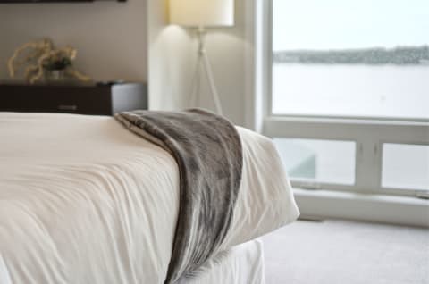 A close-up of a bed with a gray faux fur throw, beside a window showing a lake view.