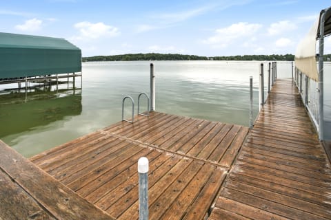 Wooden dock leading into calm lake waters with a boat house nearby and a green tree line in the background.