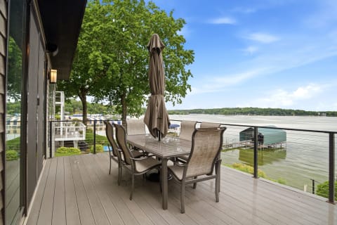 A waterfront deck with a large table and umbrellas overlooking a peaceful lake, surrounded by greenery.