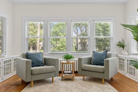 A bright sitting area with two green armchairs, a wooden side table with a potted plant, and large windows.