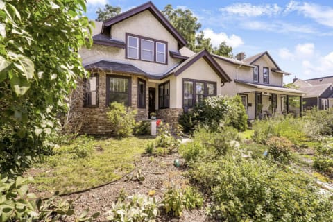 A two-story house surrounded by vibrant gardens and greenery under a blue sky.