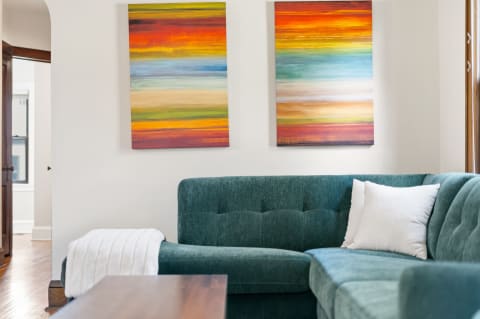 A cozy living area featuring a teal sectional couch, a white pillow, and two colorful abstract paintings on the wall.