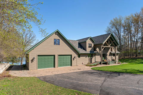 A modern tan house with green accents and a cobblestone driveway in a lush setting.
