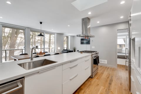 A spacious modern kitchen with a white island, wooden floors, and large windows overlooking nature.