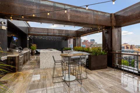 A modern rooftop terrace featuring BBQ grills, a bar counter, and a stunning city skyline view, adorned with string lights and lush plants.