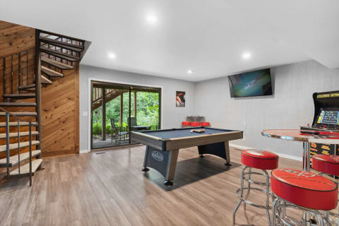 A game room with a pool table and arcade game, featuring a view of greenery through sliding glass doors.