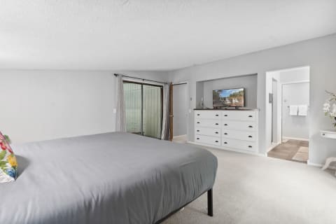 A spacious bedroom featuring a grey bedspread, colorful pillow, and modern furniture.