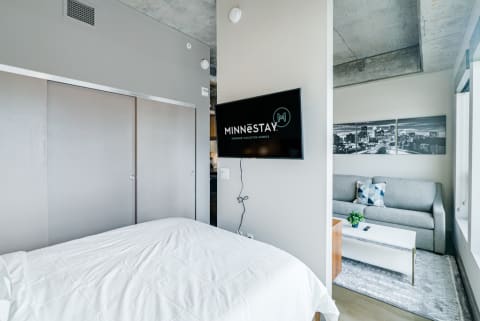 A cozy modern bedroom and living area with a white bed and gray sofa, featuring a mounted TV with MINNESTAY logo.