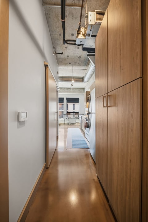 A narrow hallway with exposed concrete ceilings and sleek wood cabinetry, leading to a modern kitchen with large windows.