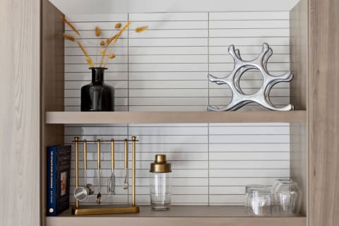A modern shelving unit with a black vase, golden dried flowers, silver sculpture, golden cocktail tools, and glassware, set against a tiled background.