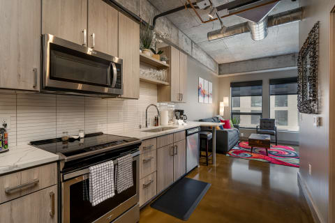 A contemporary kitchen with light wood cabinets and a cozy living area featuring a gray sofa and colorful rug.