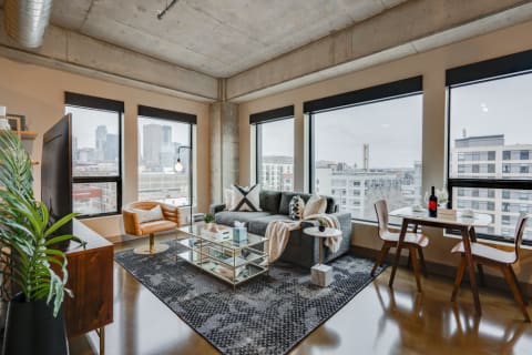 A modern living room with city skyline views, featuring a gray sectional couch, glass coffee table, and a small dining area.