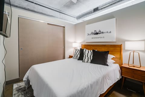 A modern bedroom with a bed, decorative pillows, a nightstand with a lamp, and a skyline artwork on the wall.