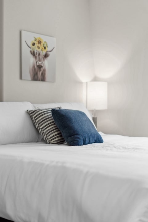 Bedroom with a white bed, decorative pillows, and a whimsical cow painting.