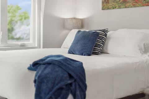 A stylish bedroom with a white bedspread, decorative pillows, a throw blanket, and a window showing greenery.