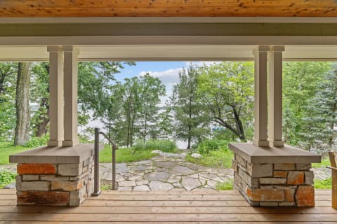 View from a porch with stone columns, featuring trees and a water body in the background.