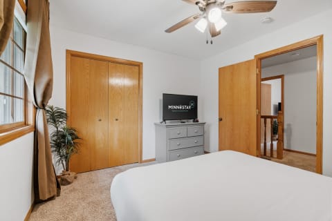 A well-furnished bedroom with a white bed, grey dresser, flat-screen TV, and natural light from a window.