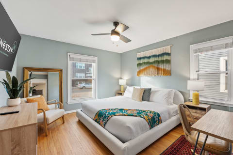 A cozy bedroom featuring a white bed with a floral throw, a macrame wall piece, and a desk.