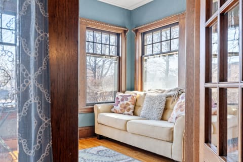 A sunlit sitting area featuring a beige couch with decorative pillows, lace curtains, and wooden window frames.